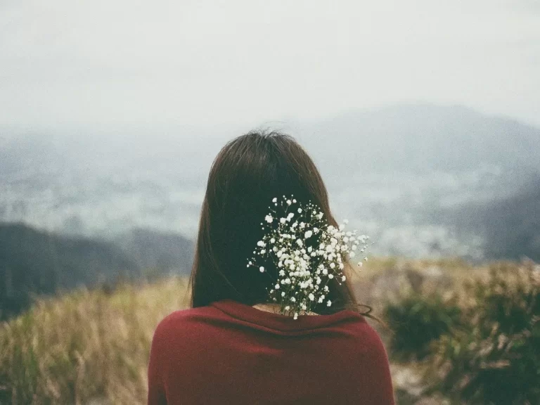 A woman in a red sweater sits outdoors, her hair adorned with delicate white flowers, overlooking a misty mountain landscape