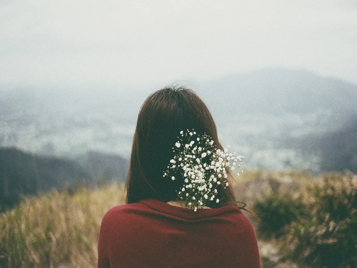 A woman in a red sweater sits outdoors, her hair adorned with delicate white flowers, overlooking a misty mountain landscape