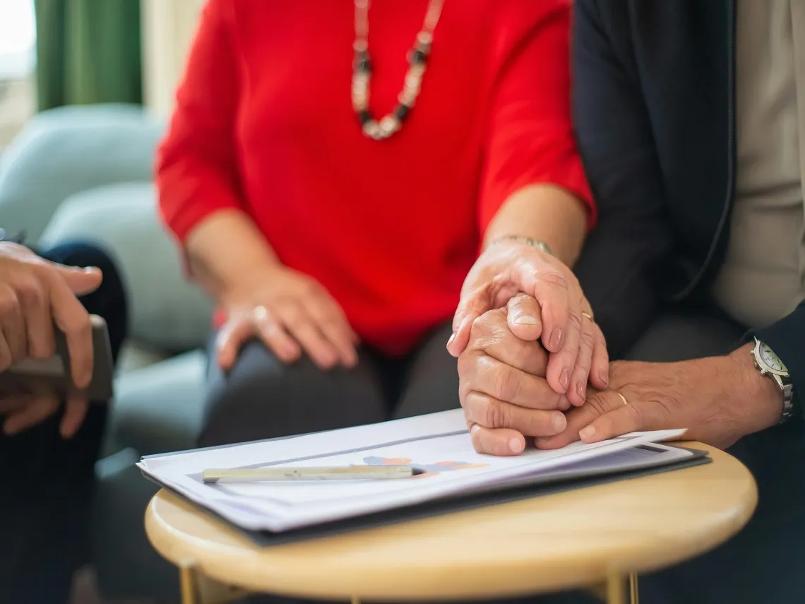 Two hands clasped together on a table with documents and a pen, symbolizing support and collaboration during a marriage counselling discussion.